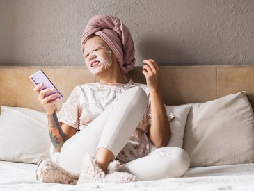 Woman relaxing in bed with face mask, towel on head and using smartphone.