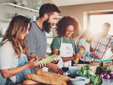 Good friends laughing and talking while preparing meals at table full of vegetables and pasta ready for cooking in kitchen