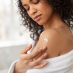 Skin care products. Young black lady applying moisturizing cream or lotion on body after shower, wearing bathrobe in bathroom, closeup, cropped