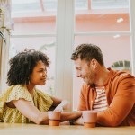 A heterosexual couple, perhaps in the honeymoon phase of their relationship look very devoted and in love as they gaze at each other at a dining table. The man looks a little shy as he momentarily breaks eye contact and looks at the ground while smiling. She touches his arm to reassure him.