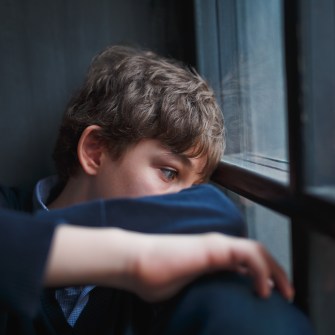 Pensive sad boy teenager with blue eyes in a blue shirt and jeans sitting at the window and closes his face with his hands.