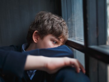 Pensive sad boy teenager with blue eyes in a blue shirt and jeans sitting at the window and closes his face with his hands.