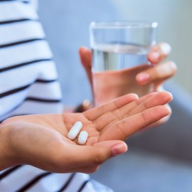 Woman holding white pill on hand and drinking water in glass on sofa in house, feels like sick. Health care concept.