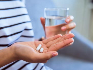 Woman holding white pill on hand and drinking water in glass on sofa in house, feels like sick. Health care concept.